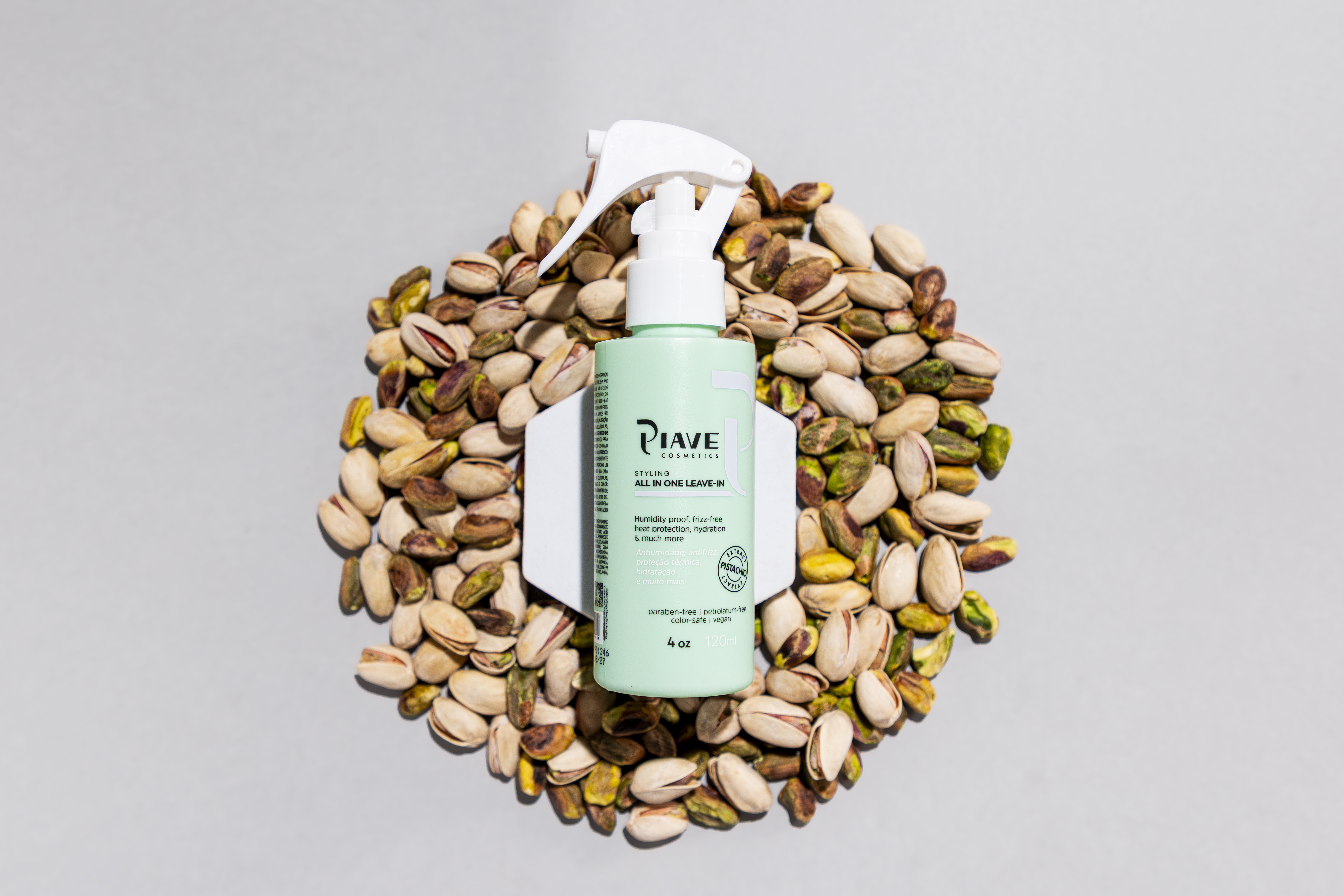 Frasco spray verde da Piave All In One Leave-In centralizado sobre um círculo de pistaches abertos, sobre fundo cinza claro